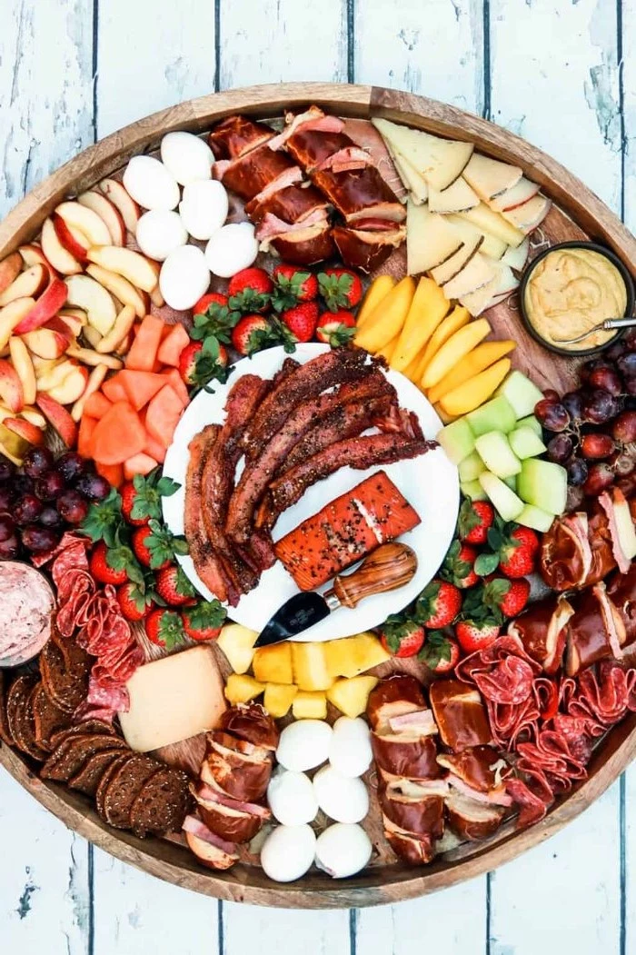 different types of cheese fruits and meet arranged in round charcuterie board made of wood