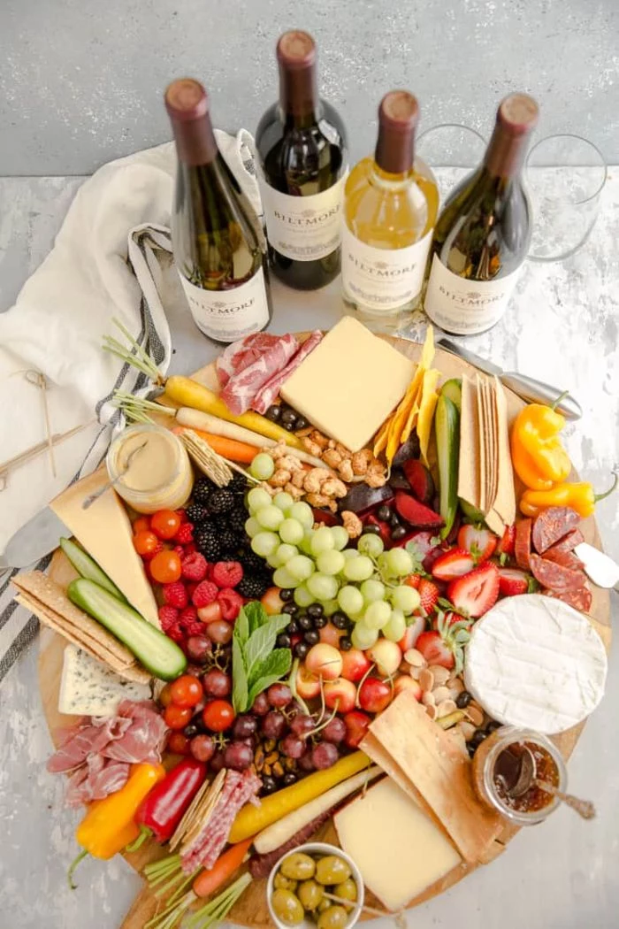 four bottles of wine next to round wooden board charcuterie board cheese meats fruits veggies crackers nuts