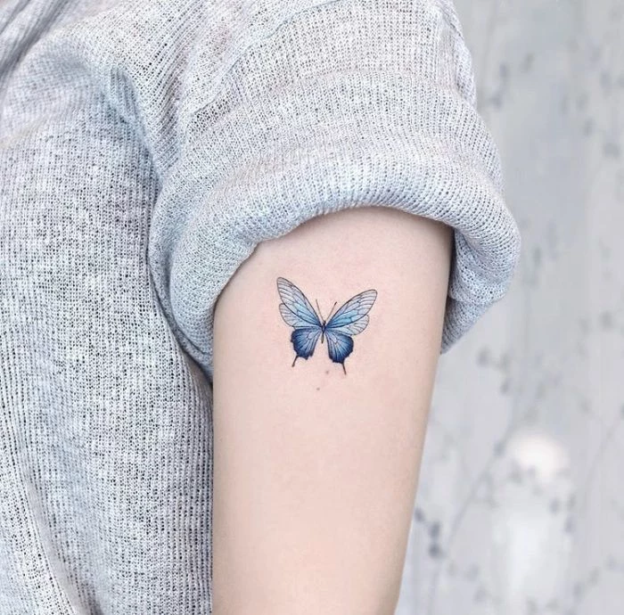 small blue butterfly on the side of the arm butterfly tattoos on shoulder on woman wearing gray sweater