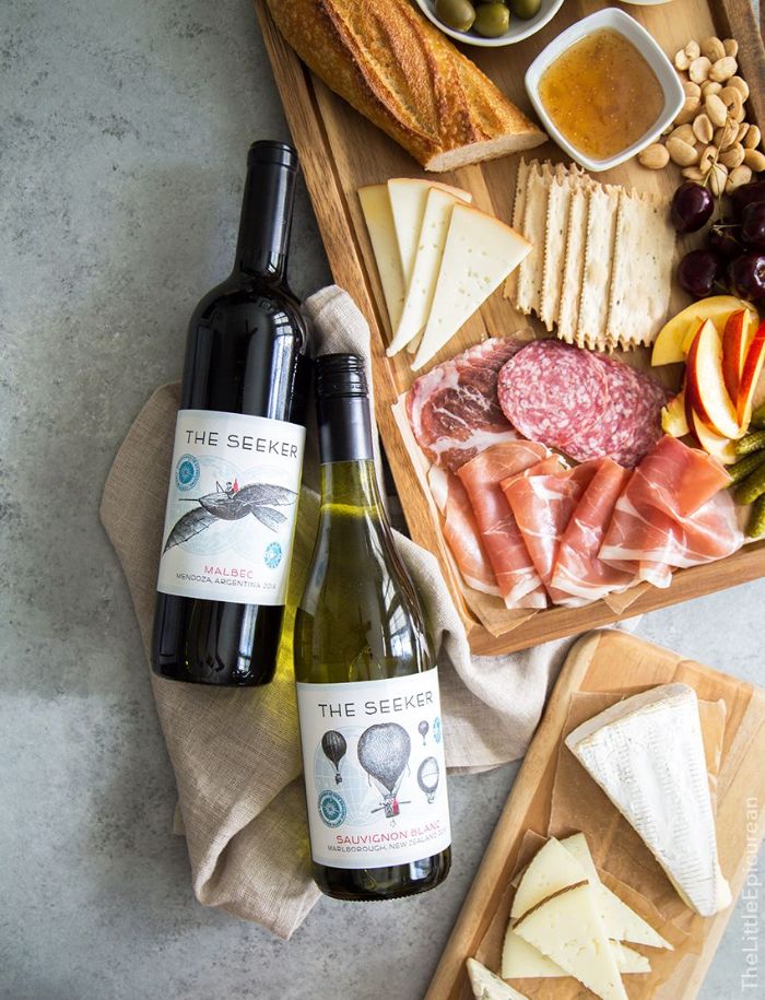 two bottles of wine placed next to wooden board meat and cheese board with baguette crackers fruits nuts