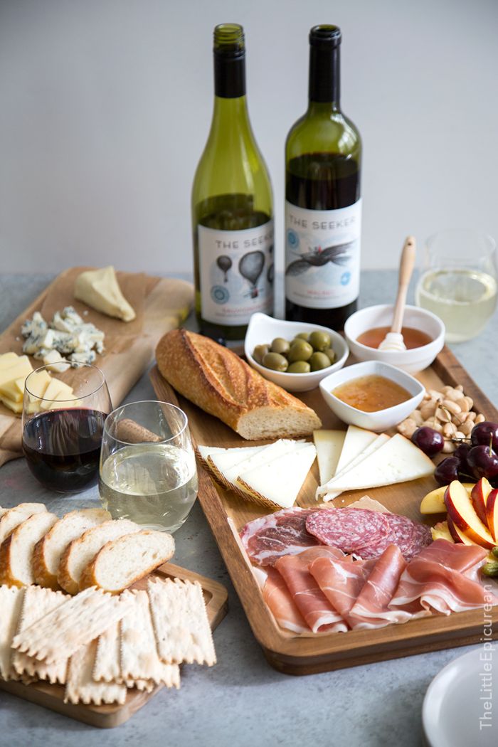 two wine glasses next to wooden board meat and cheese board baguette olives fruits and nuts