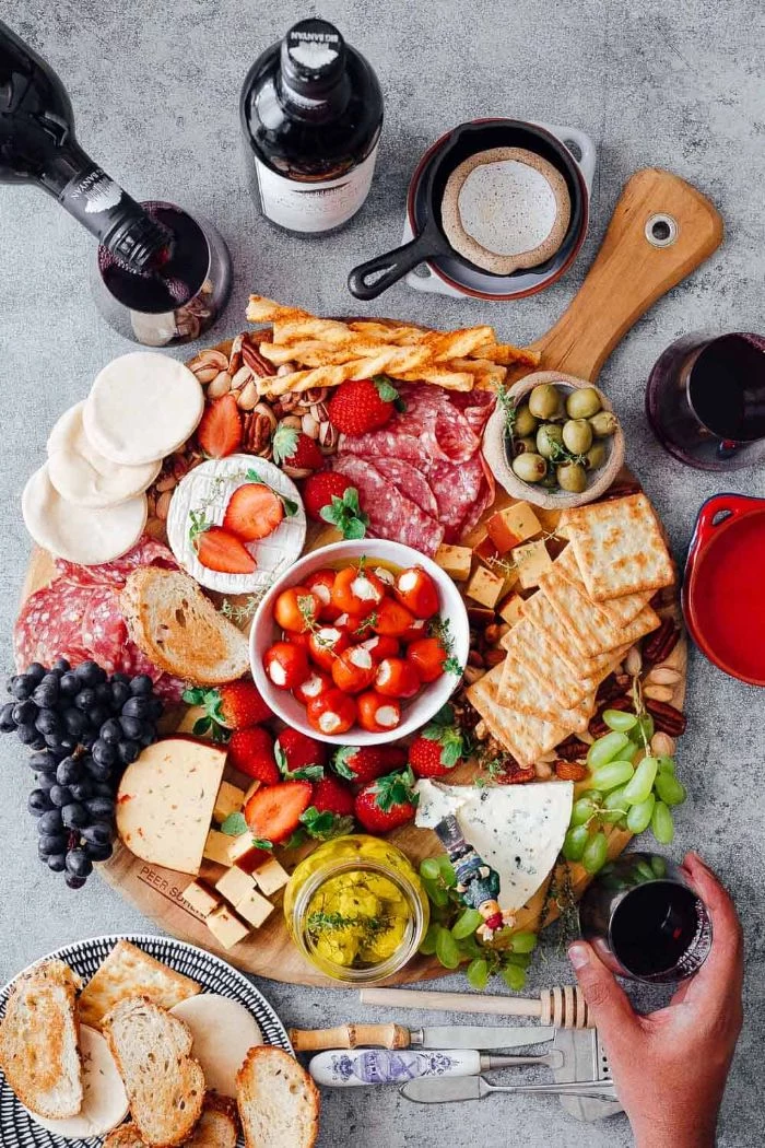 wine bottles and glass next to round wooden board meat and cheese platter with fruits veggies crackers nuts