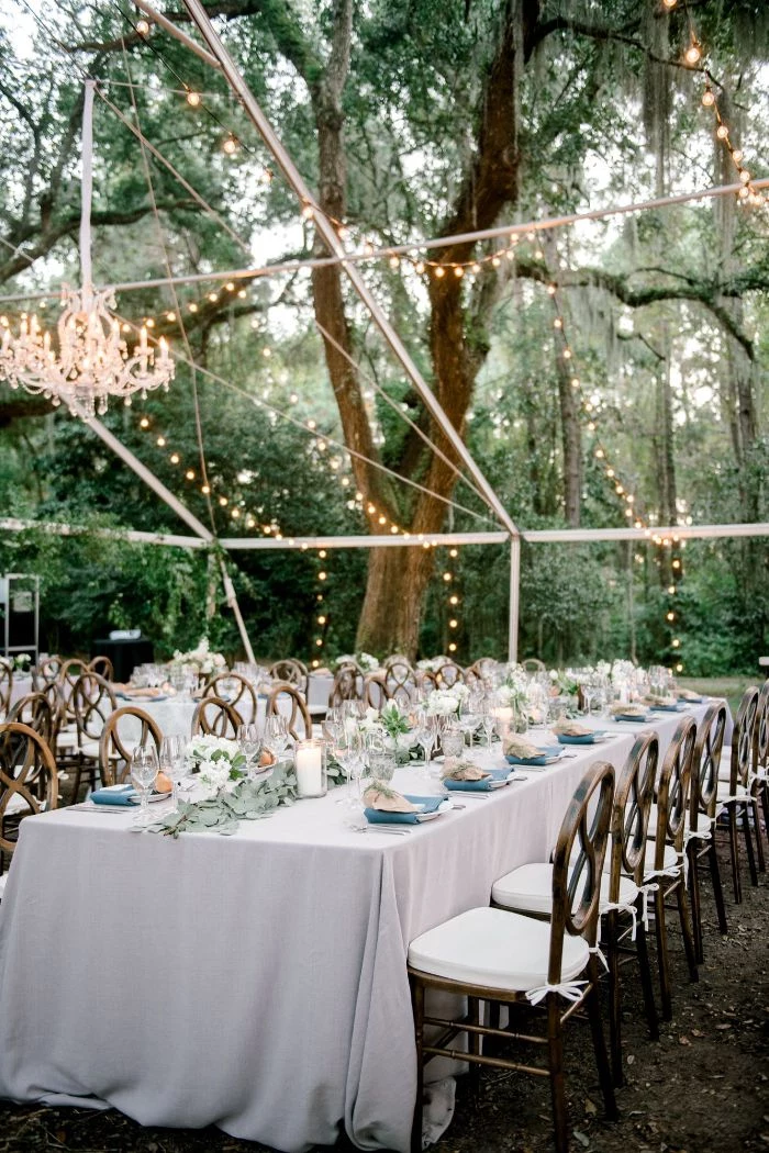 arrangements of flowers in the middle of long table with white table cloth rustic wedding ideas strings of lights hanging above