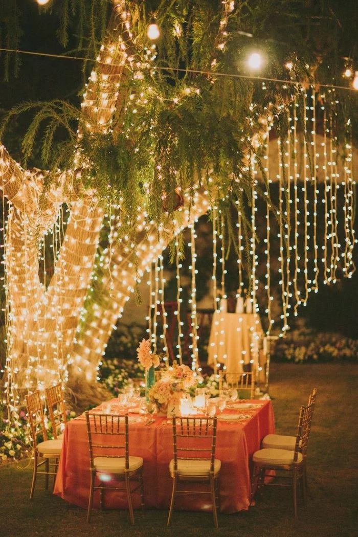 backyard wedding decorations tree wrapped with strings of lights table underneath with floral centerpiece