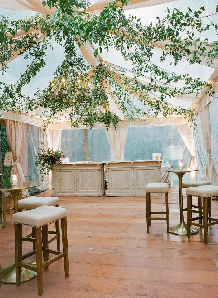 bar stools next to high tables under tent made of white curtains outdoor wedding decorations greenery hanging from the top