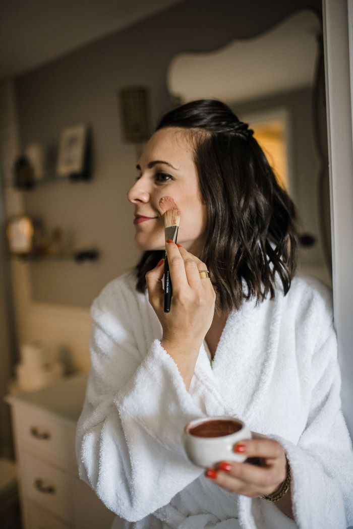 black haired woman wearing bath robe skin care masks putting on mask on her face with brush