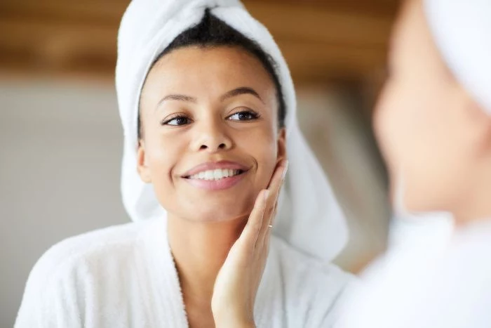 black haired woman wearing white bath robe and towel on her head hydrating face mask looking at a mirror