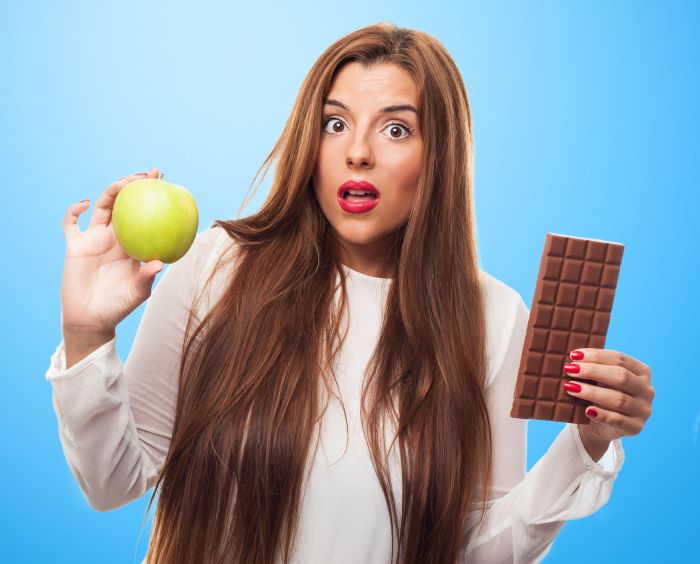 brunette woman wearing white blouse standing on blue background holding an apple and chocolate food education