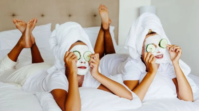 two women wearing white face masks with two cucumber slices on their eyes honey face mask