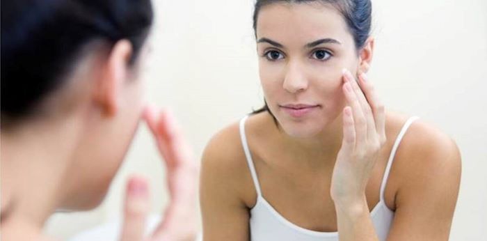 woman looking at herself in the mirror homemade face mask for acne with black hair wearing white top