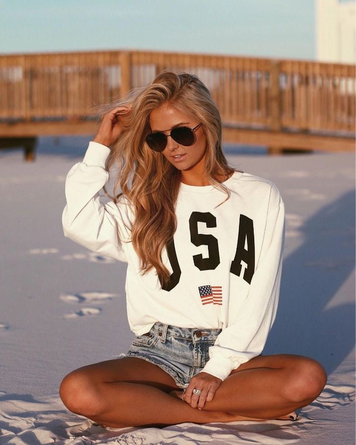blonde girl sitting on the beach clothes for teenage girls wearing denim shorts white long sleeved usa blouse