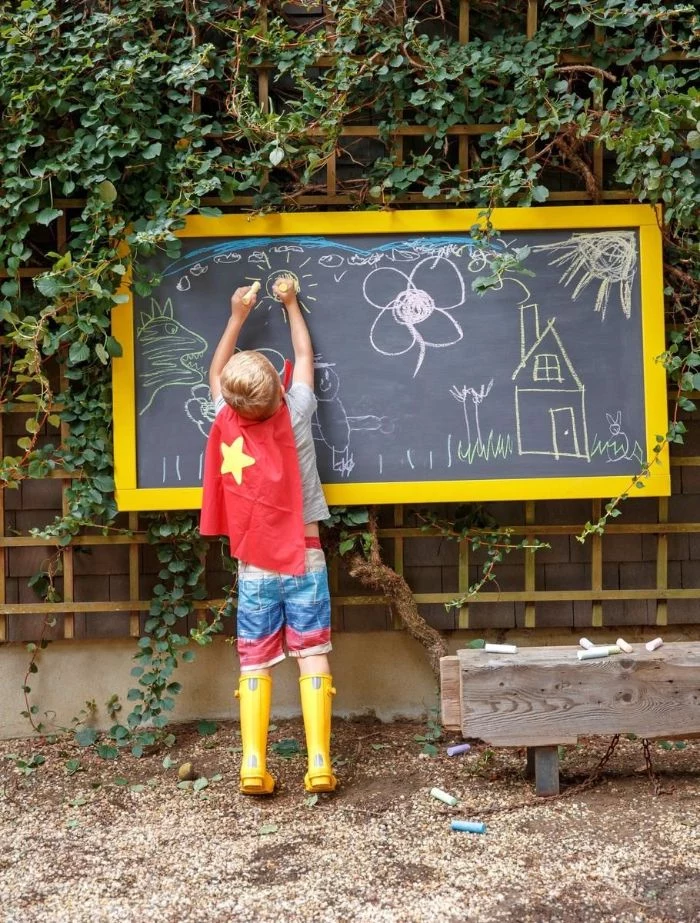 boy wearing superhero cape yellow boots drawing on a chalk board games to play outside