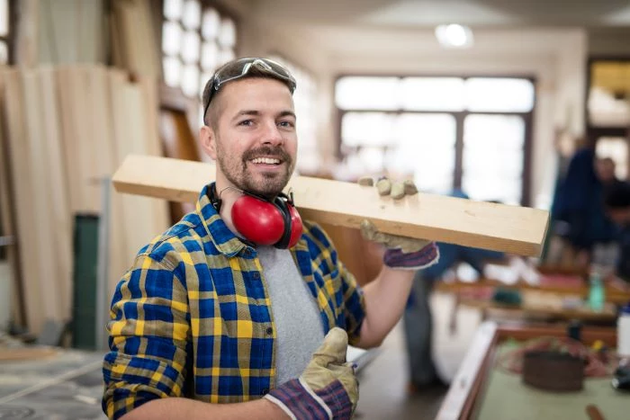 happy smiling carpenter holding wood material home remodeling wearing blue yellow plaid shirt goggles