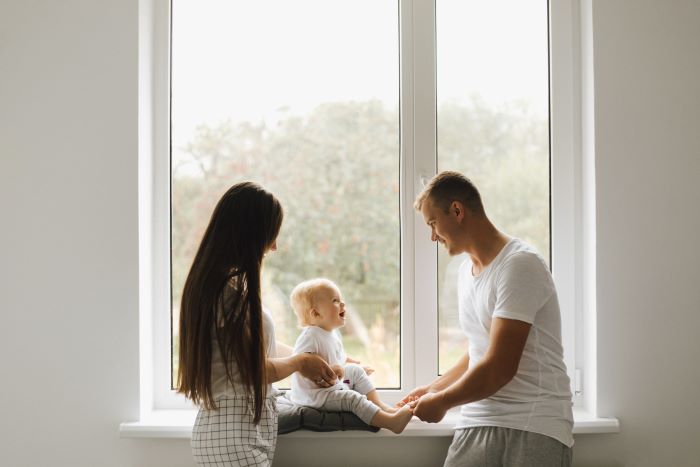 mom dad play with their little son sitting on window frame in front of large window installation