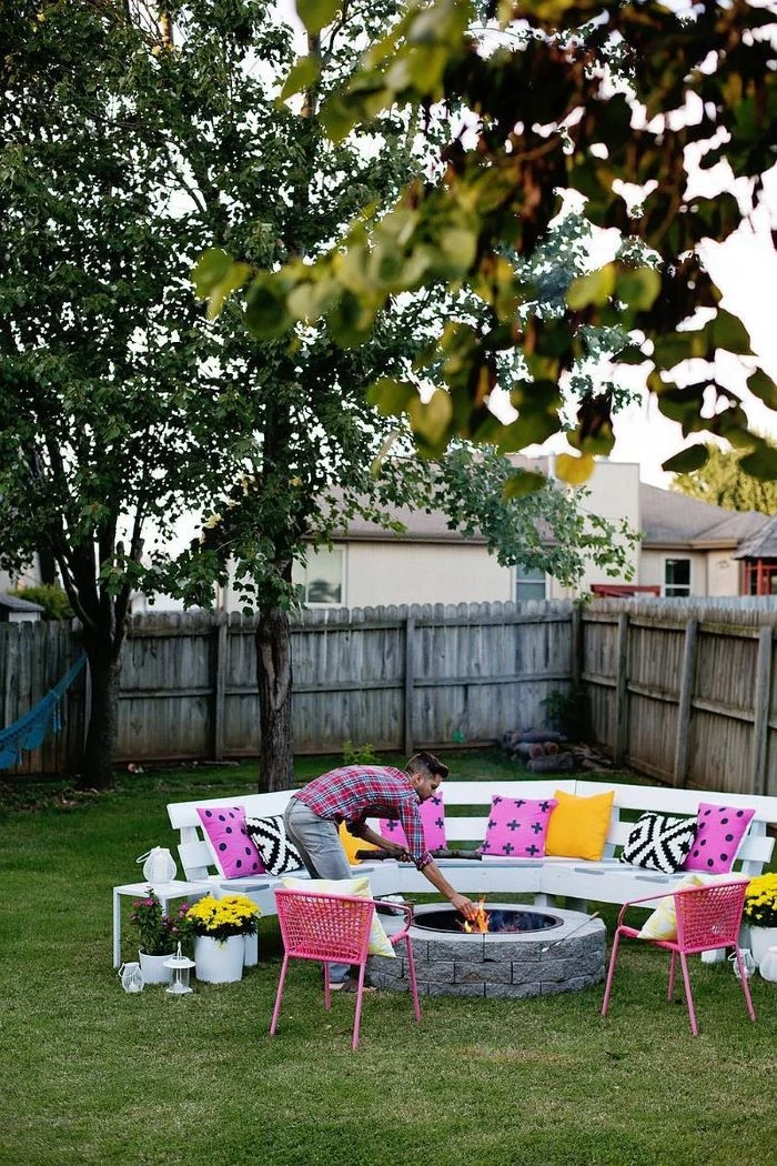 white wooden bench with colorful throw pillows build your own fire pit with stones man throwing logs into the fire