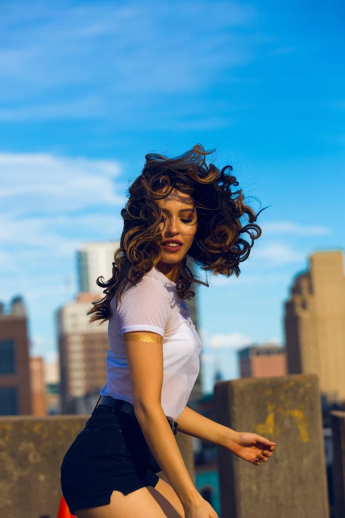 brunette woman with wavy hair wearing white t shirt black shorts diy hair mask standing on top of building