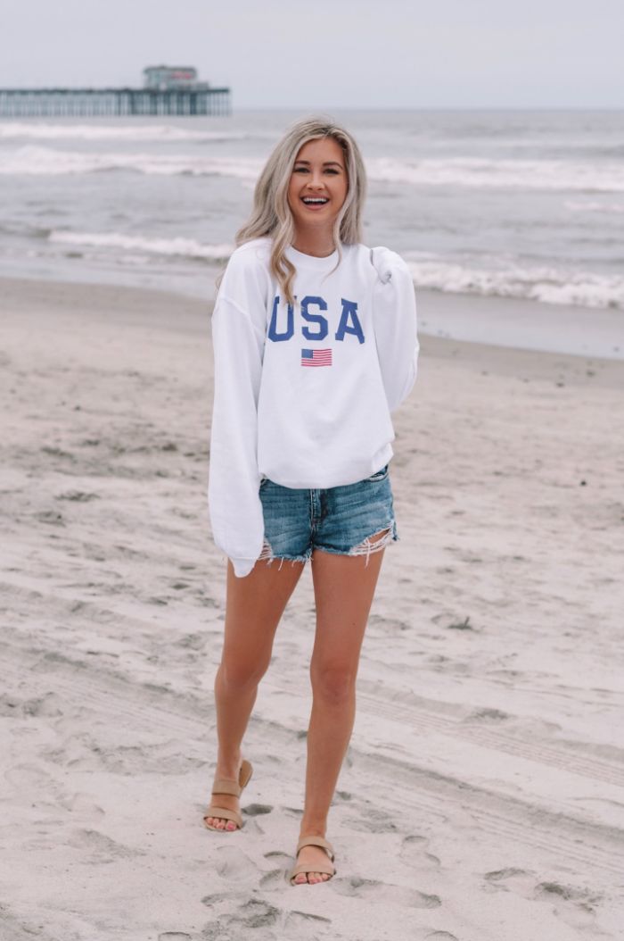denim shorts white usa sweatshirt worn by woman with long blonde hair 4th of july clothes on the beach
