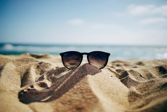 ray ban sunglasses placed on the sand beach aesthetic blurry sky and ocean in the background