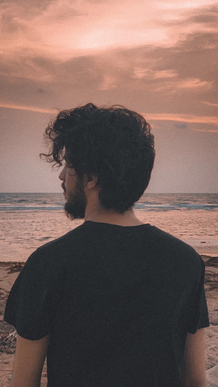 waves in the background best hair mask for damaged hair man standing on the beach with curly black hair wearing black t shirt