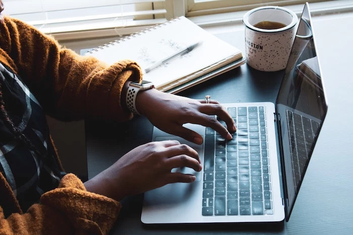 woman wearing orange sweater typing on laptop keyboard working from home coffee mug next to her