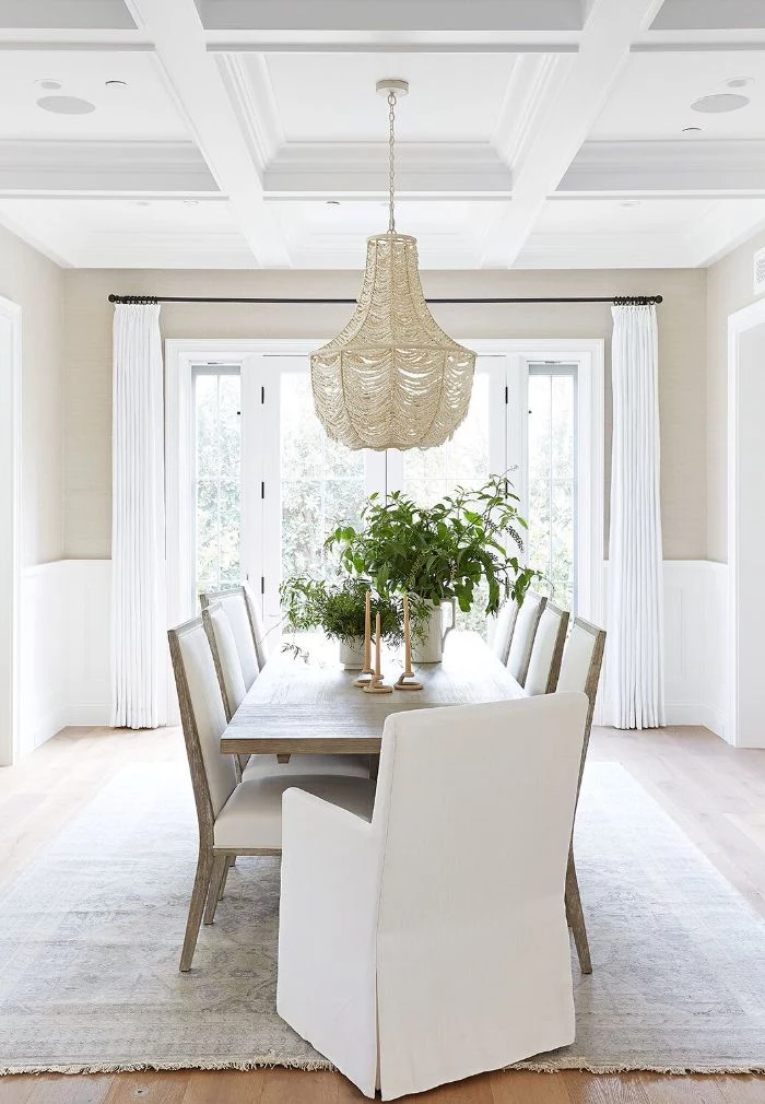 chandelier hanging above long rustic dining table white wooden chairs around it tall windows