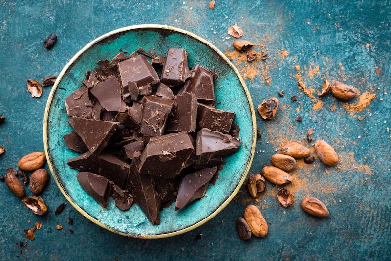 pieces of broken dark chocolate in a turquoise bowl on blue stone table with cocoa beans and powder