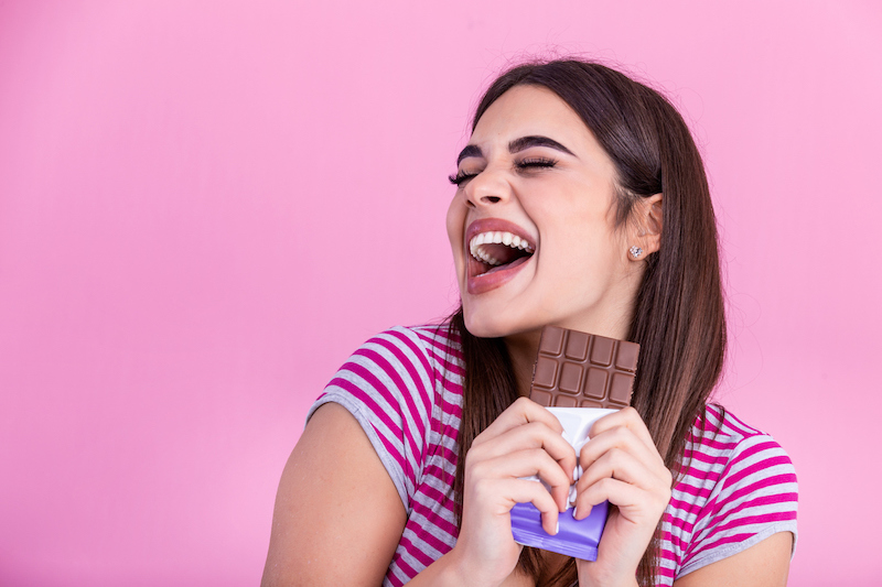 happy young beautiful lady eating chocolate and smiling. girl tasting sweet chocolate. young woman with natural make up having fun and eating chocolate isolated on pink background