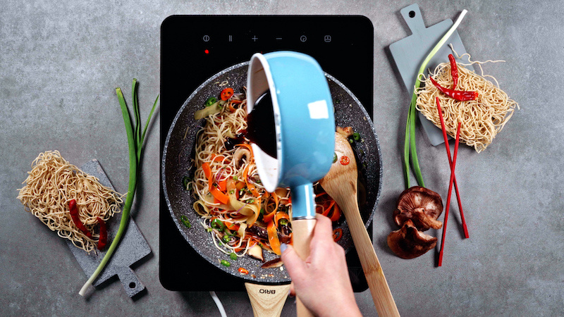 sweet and sour sauce being poured into stir fry of veggies and noodles