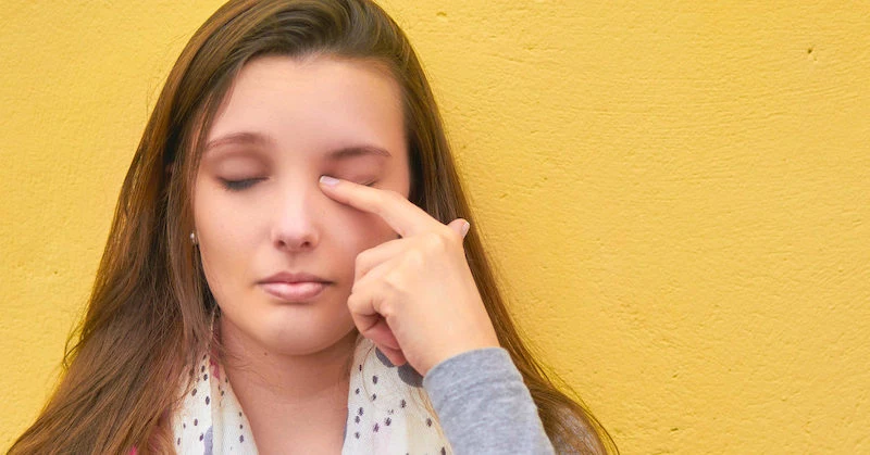 young woman rubbing eye while standing against yellow wall