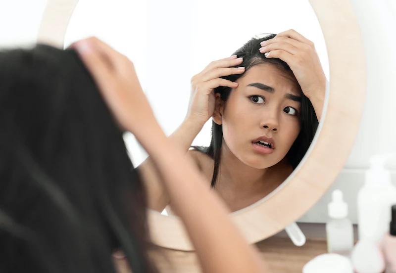 worried asian woman checking her hair in mirror at home