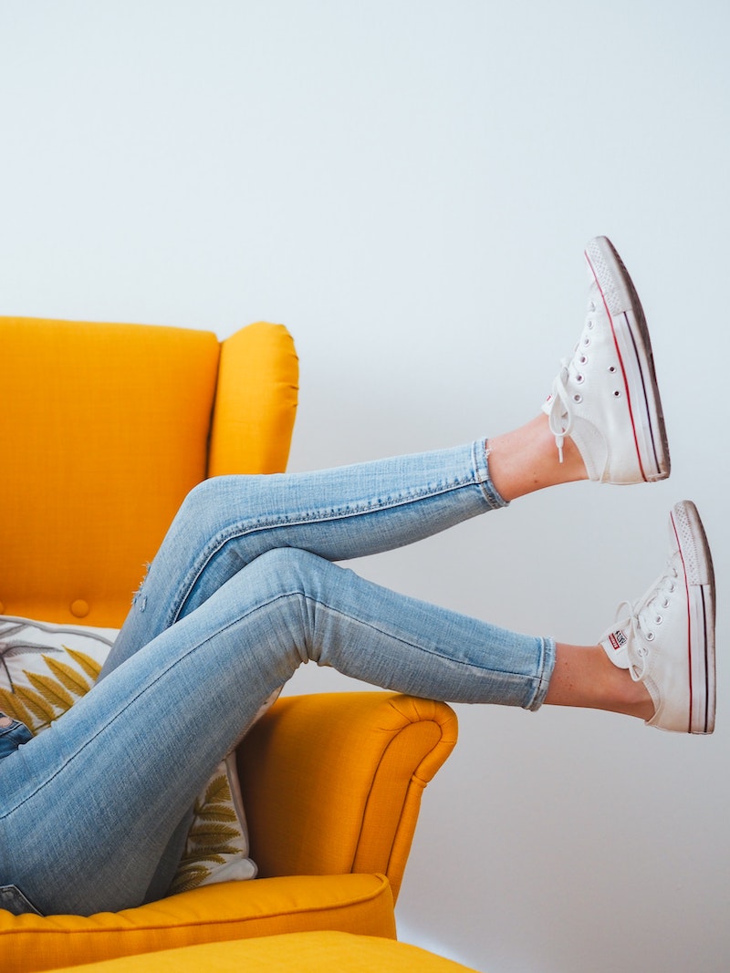 woman sitting sideways on yellow couch with legs up in jeans and white converse