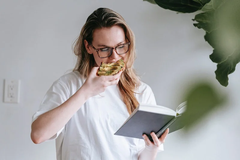 woman eating avocado toast while reading a book